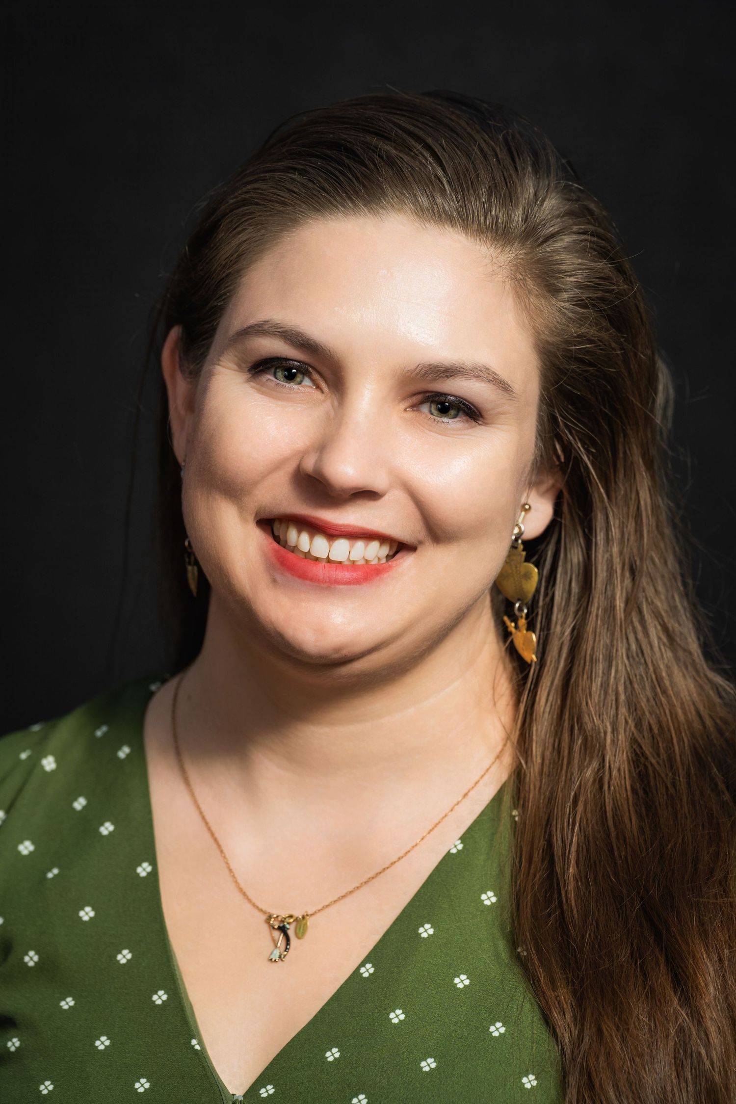 Professional headshot with person wearing green polka dot top and gold jewelry against dark backdrop.