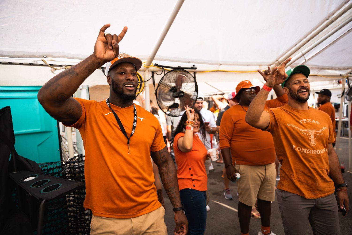 Fans in orange clothing dancing and celebrating at an outdoor event under a tent.