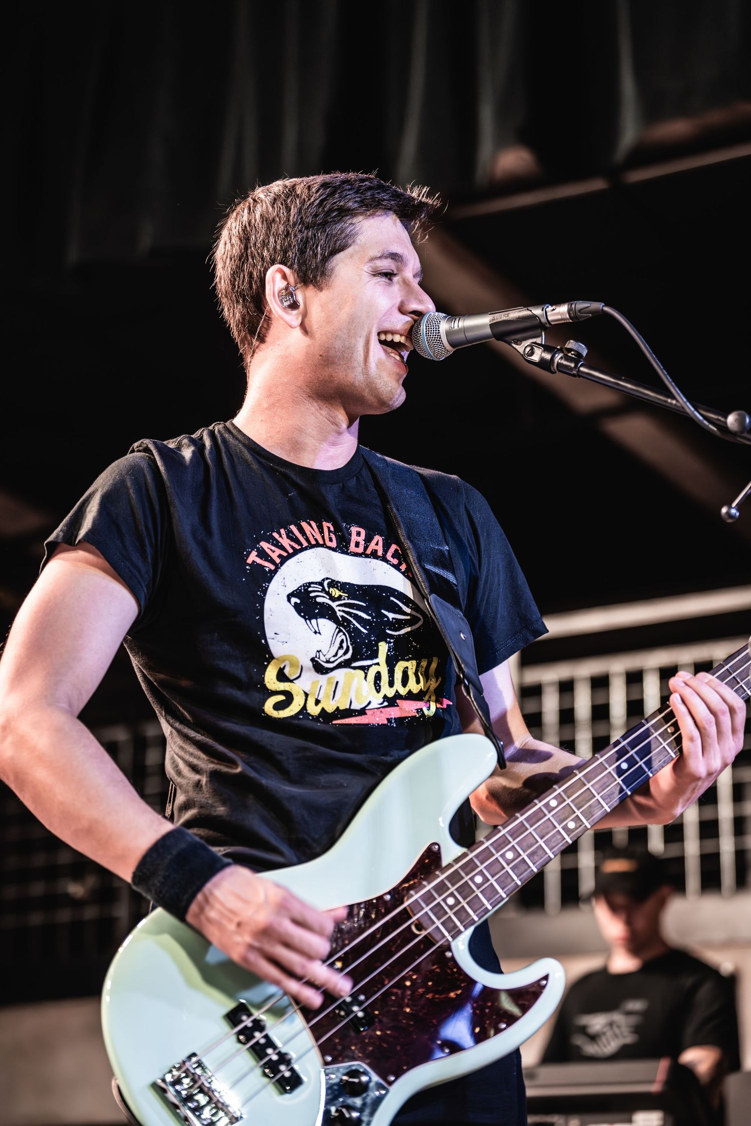 Bassist plays Fender bass guitar while singing into microphone during live performance.