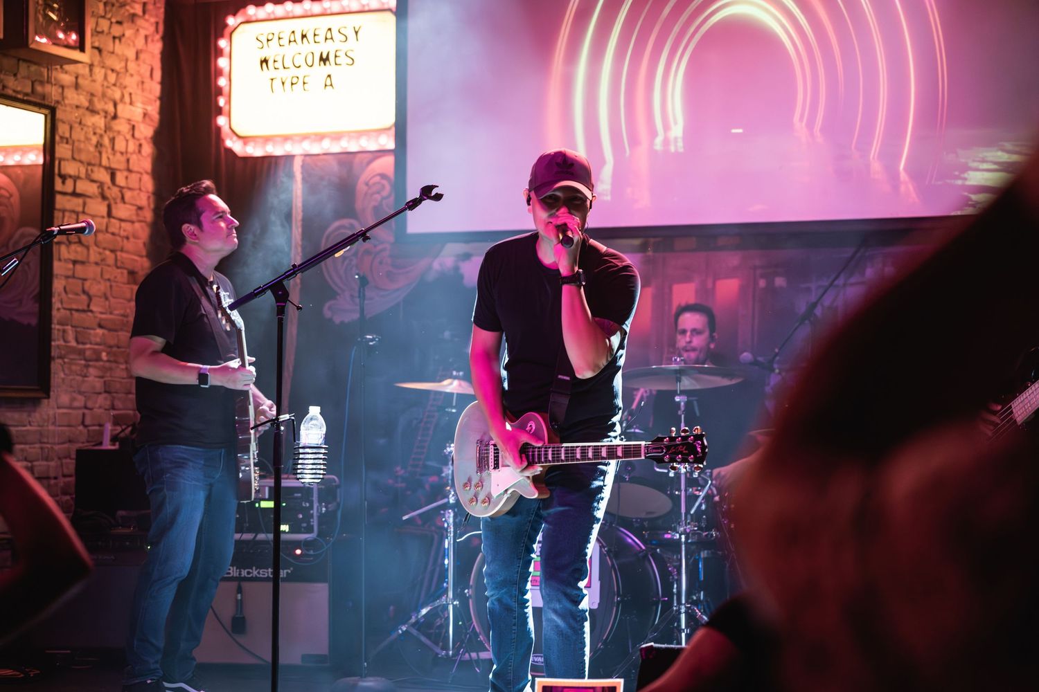 Band performing on stage with pink lighting and brick wall backdrop at intimate venue.