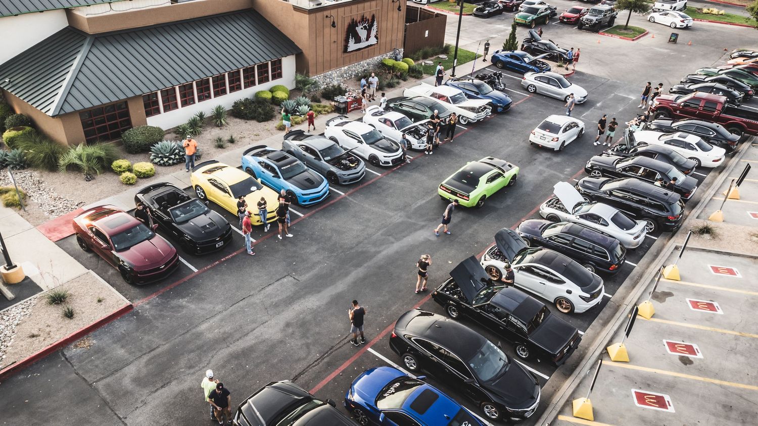 Car enthusiasts gather at a restaurant parking lot for an automotive meetup viewed from above.