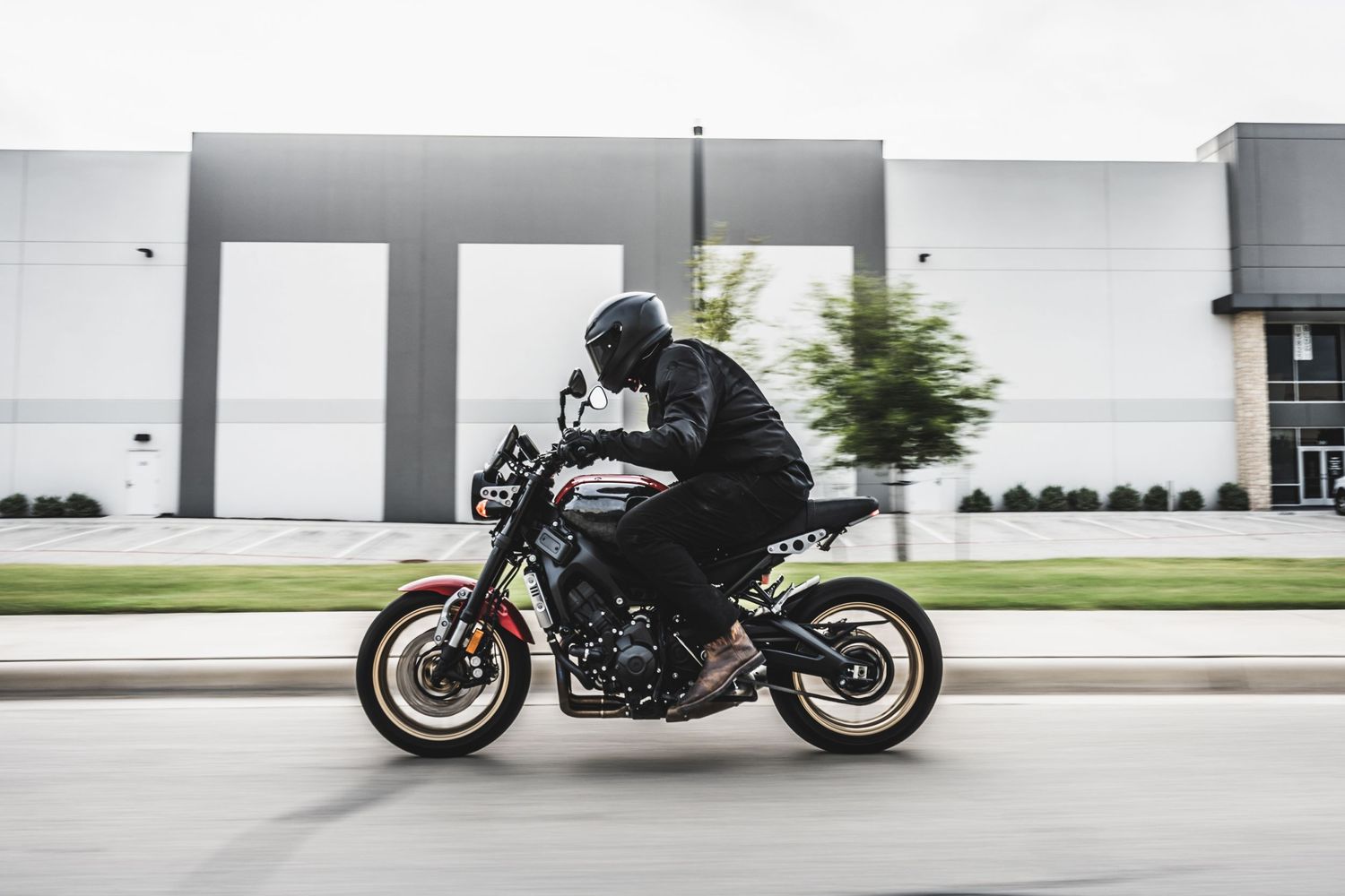 Side profile of motorcycle rider on black street bike passing industrial buildings.