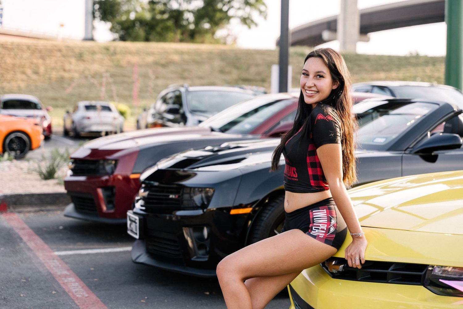 Woman in black outfit sitting on yellow sports car in outdoor dealership setting.