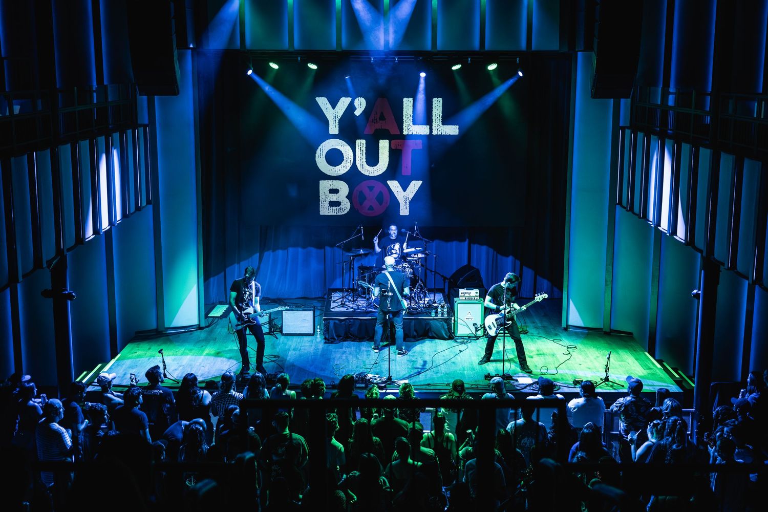 Wide shot of band performing on stage with Y'all Out Boy signage and enthusiastic audience below.