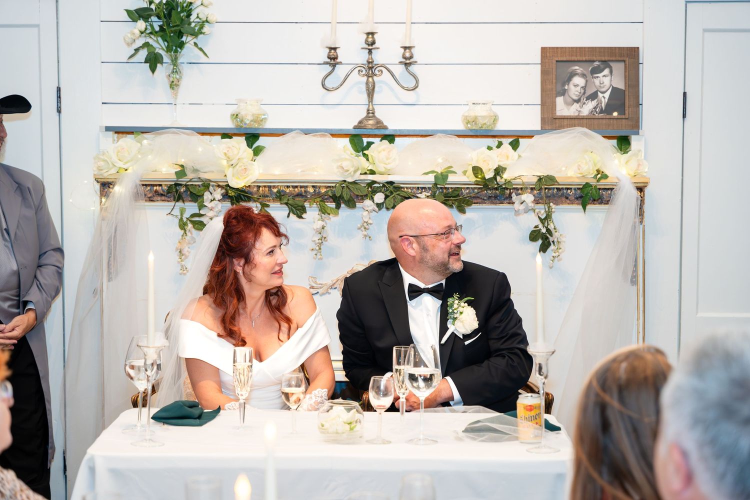 Newlyweds seated at their reception table with floral decorations and candelabras on the wall behind them.