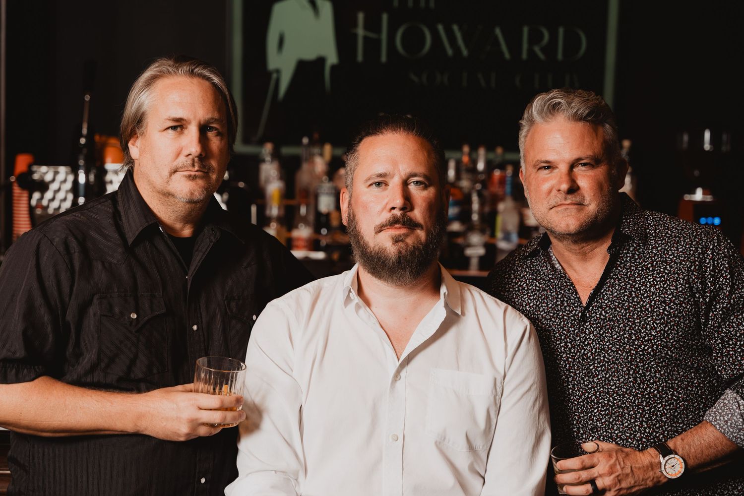 Three men in casual attire pose together at bar with dark ambient lighting.
