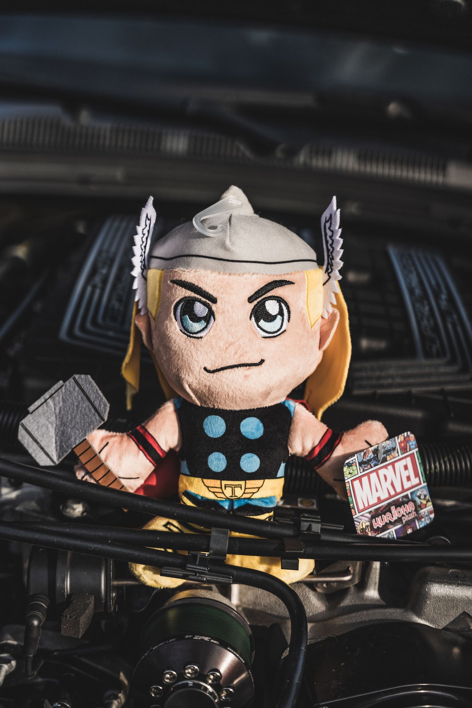 Thor plush toy sitting on car engine compartment with Marvel tag attached.