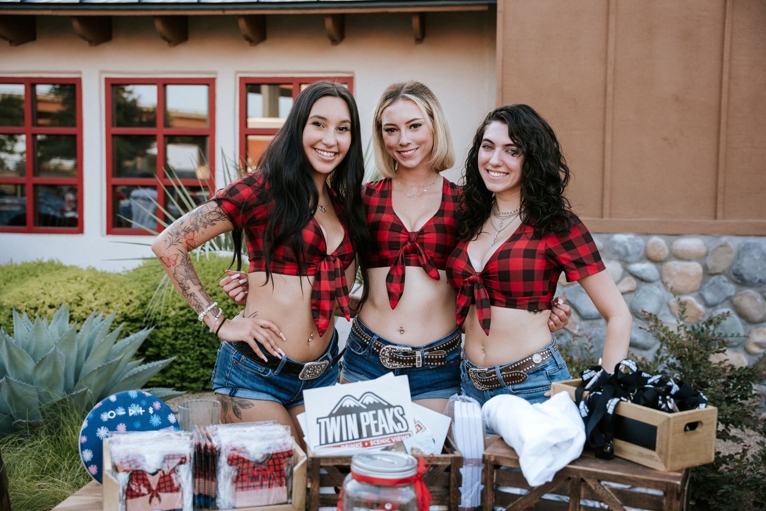 Three women in matching red plaid shirts and denim shorts posing with merchandise at an outdoor booth.