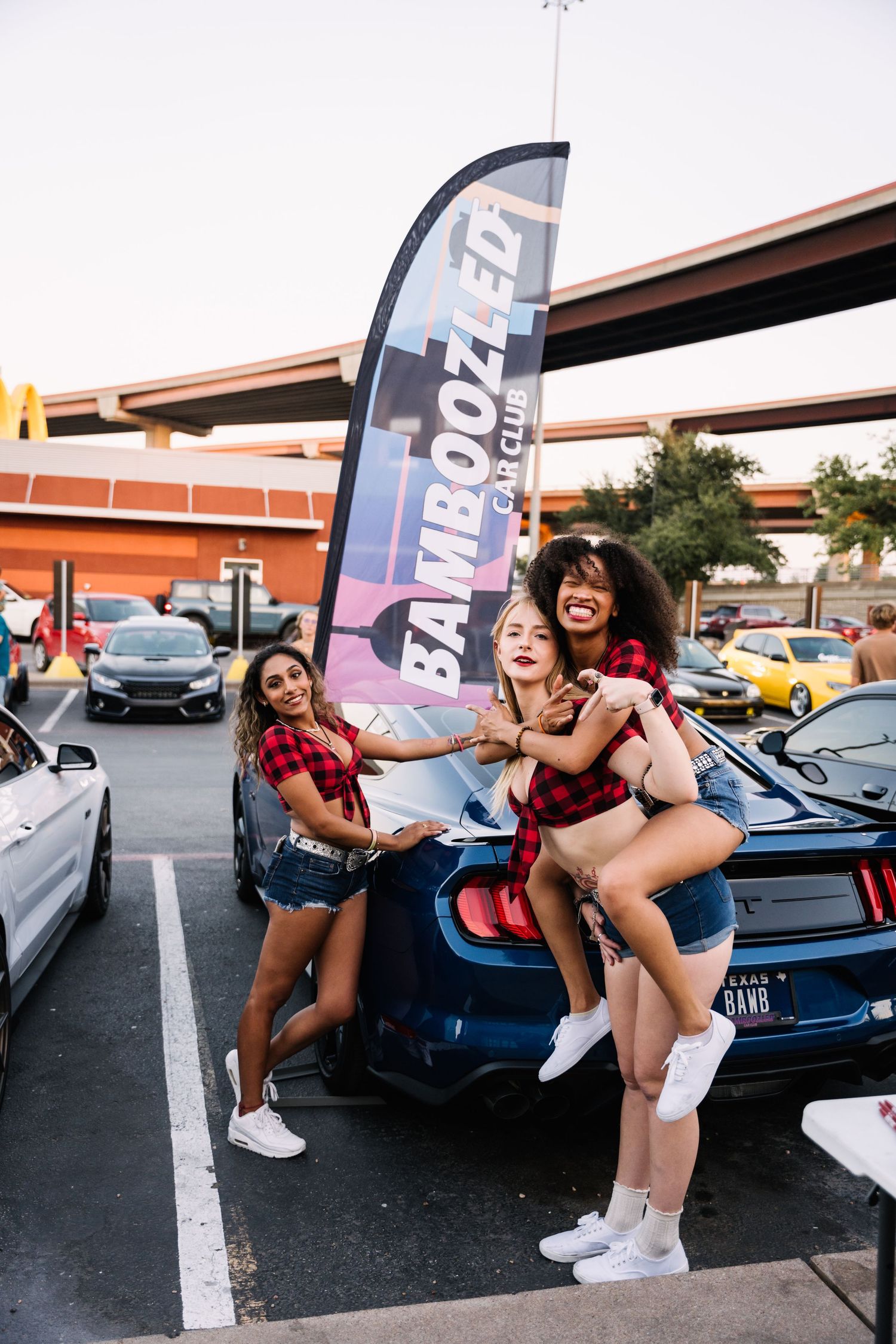 Group posing energetically next to cars at an outdoor car meet with a Fanboost banner.