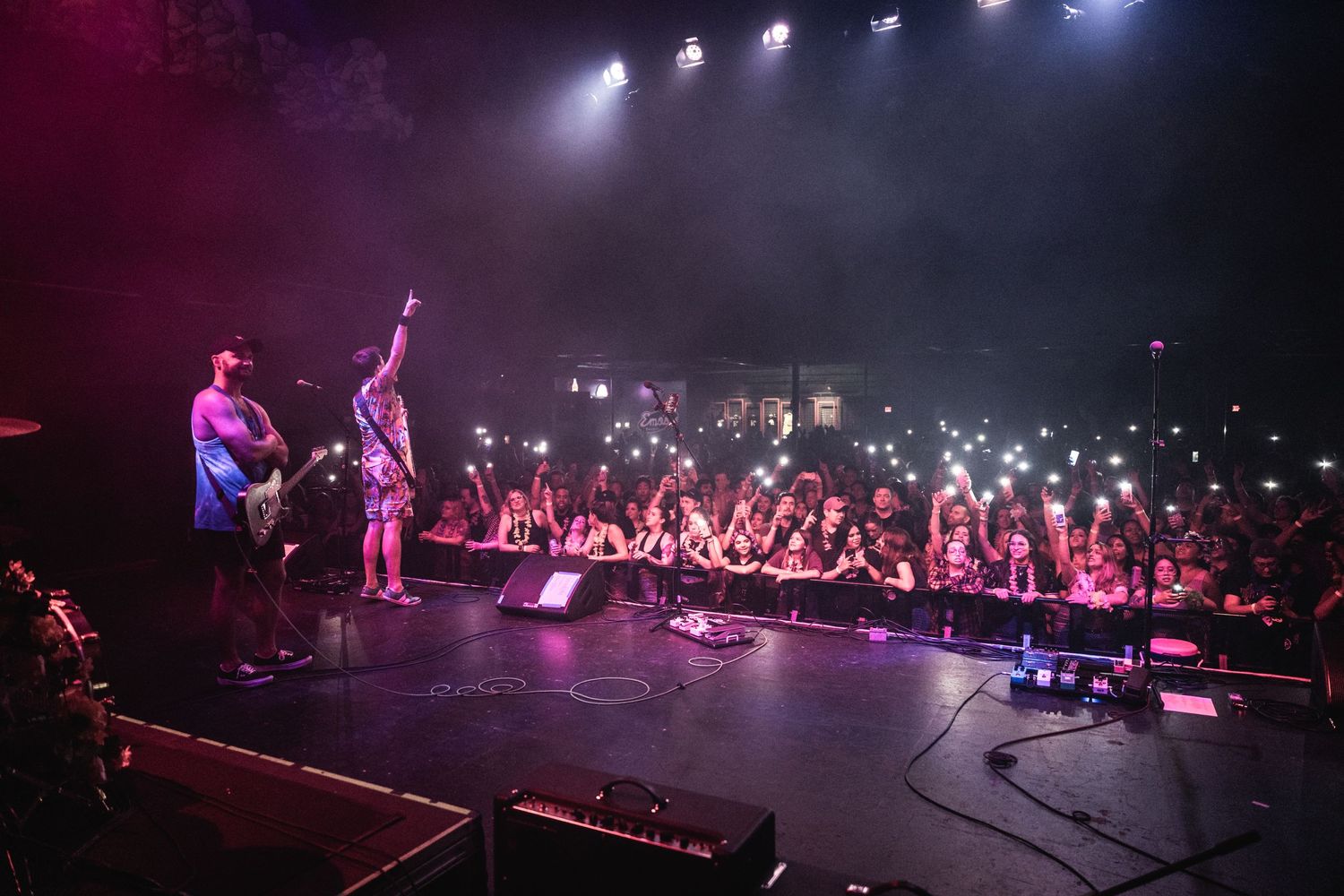 Concert venue filled with energetic crowd and stage lighting during live performance with band silhouettes.