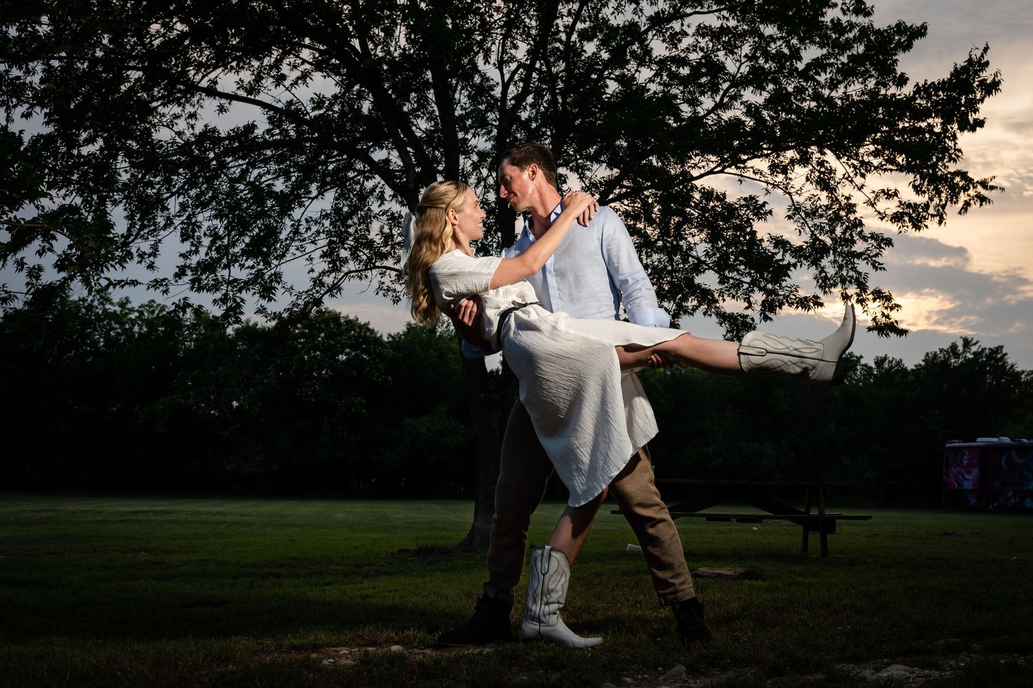 A couple performs a dramatic dance pose at dusk with trees silhouetted in the background.
