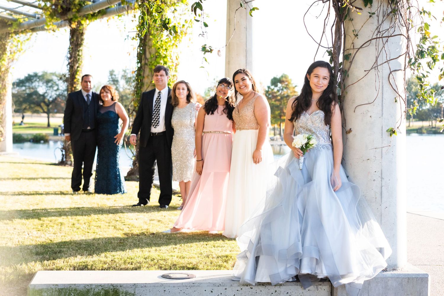 Wedding party poses outdoors in formal attire near waterfront setting.