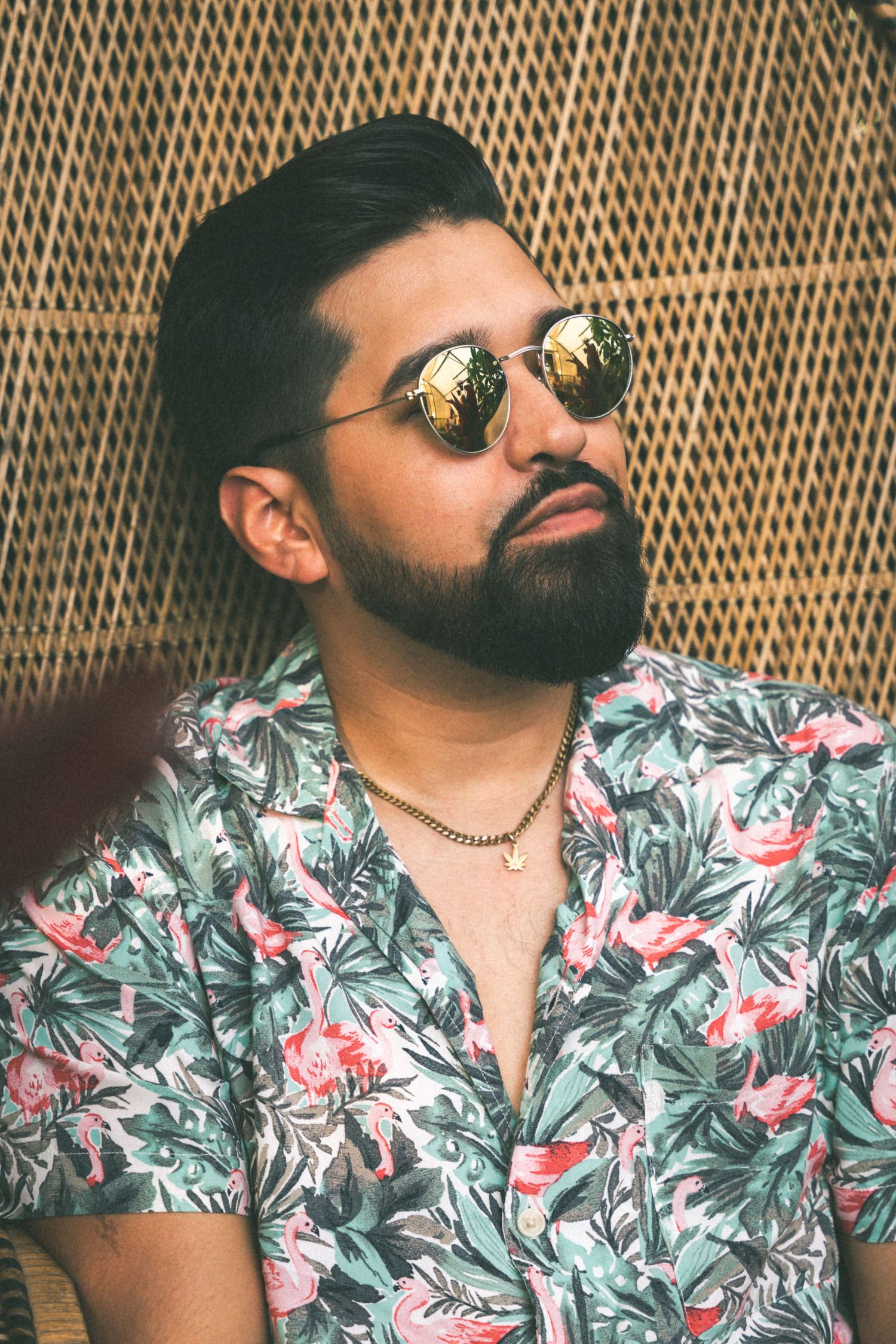 Stylish man in floral shirt and sunglasses poses against woven backdrop.