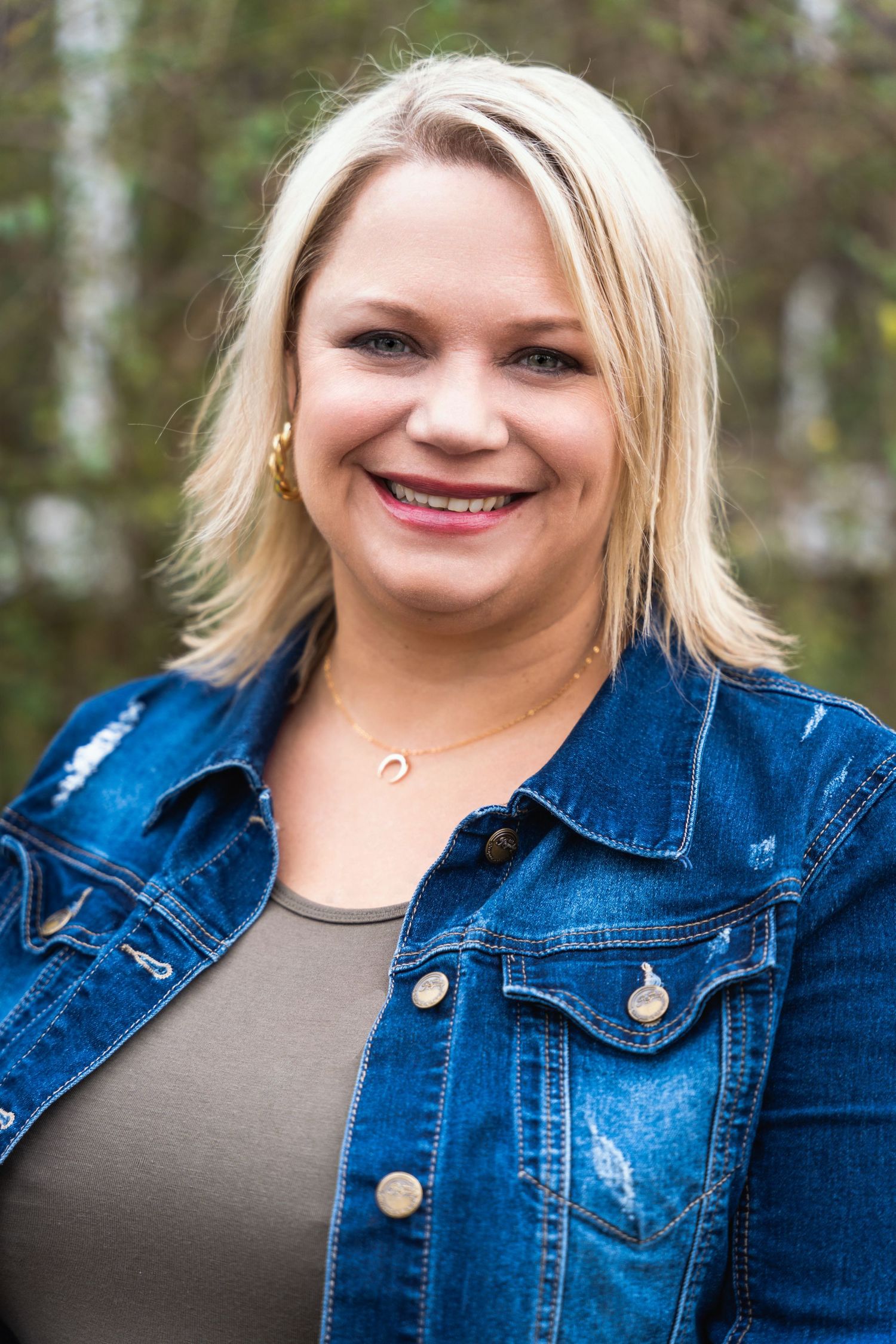 Casual portrait of person wearing denim jacket and gray top with blonde hair against trees.