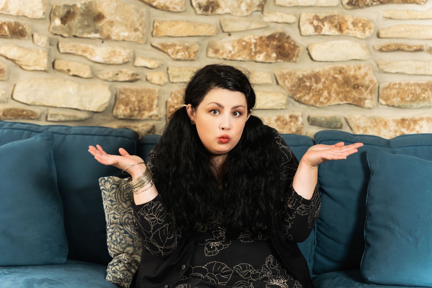 Expressive portrait on blue couch with stone wall backdrop showing questioning gesture.