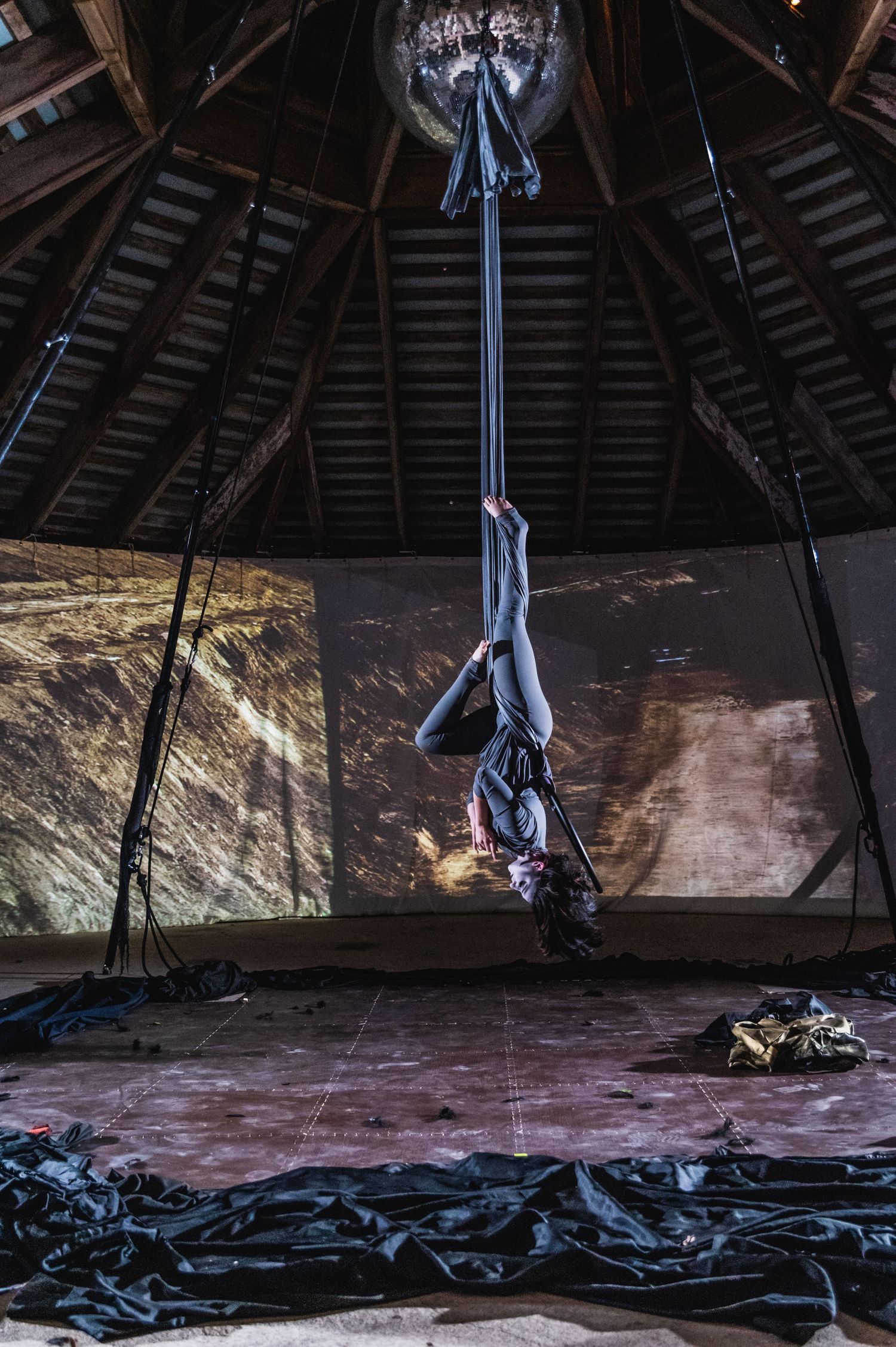 Aerial silk performer suspended in dramatic pose beneath wooden rafters.