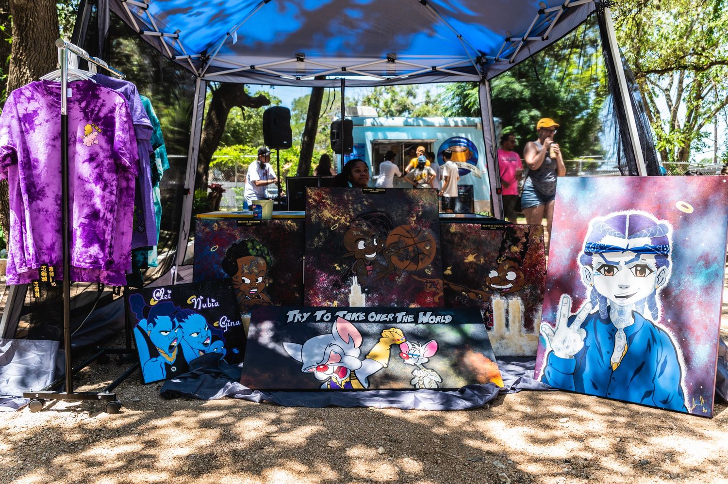A vendor booth displaying colorful anime-style artwork and merchandise under a blue canopy tent.