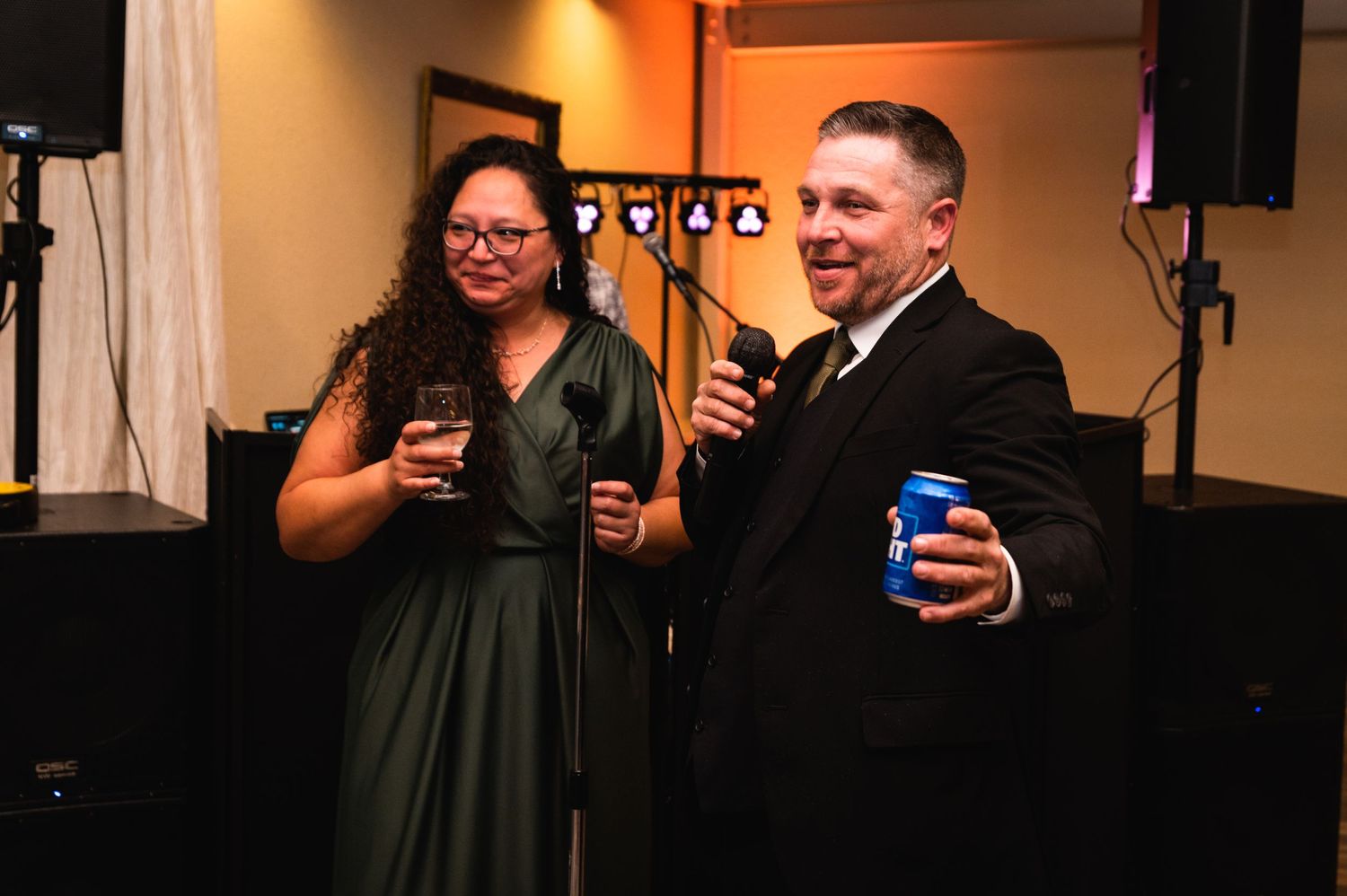 Two guests share drinks and smiles during a wedding reception celebration in a warmly lit venue.