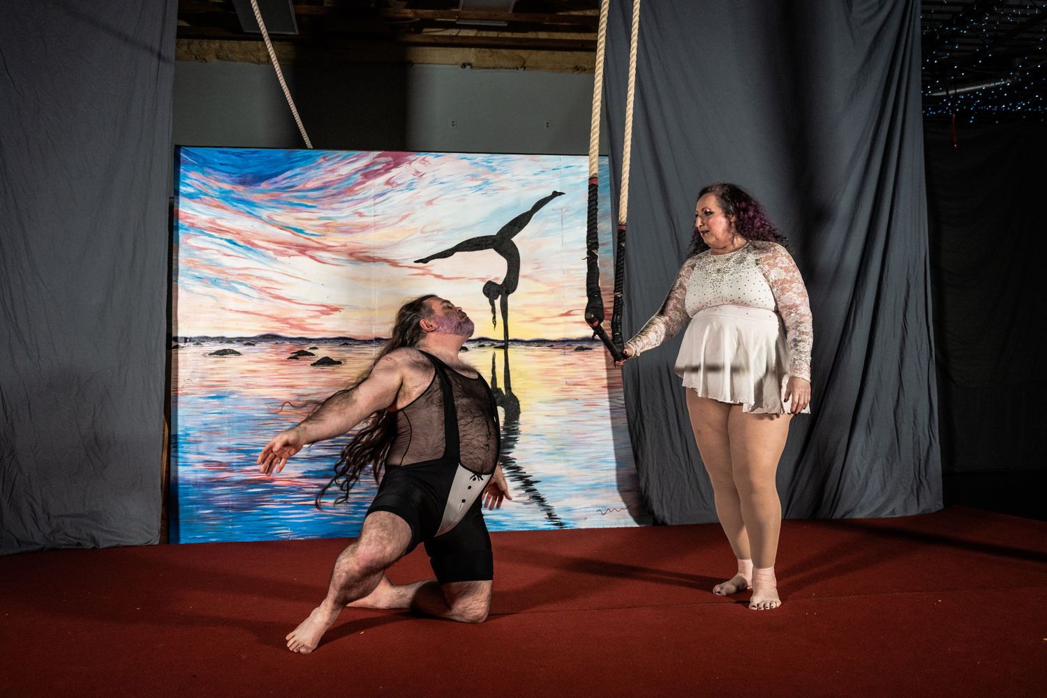 Two performers engage in an aerial dance routine against a painted sunset backdrop.