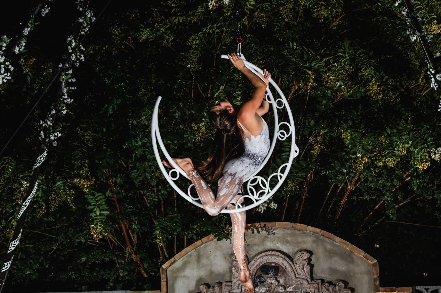 Graceful aerial dance performance on a crescent moon prop with garden backdrop.
