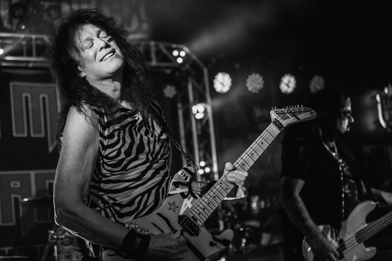 Stage performer plays electric guitar wearing zebra print tank top during dramatic concert performance.