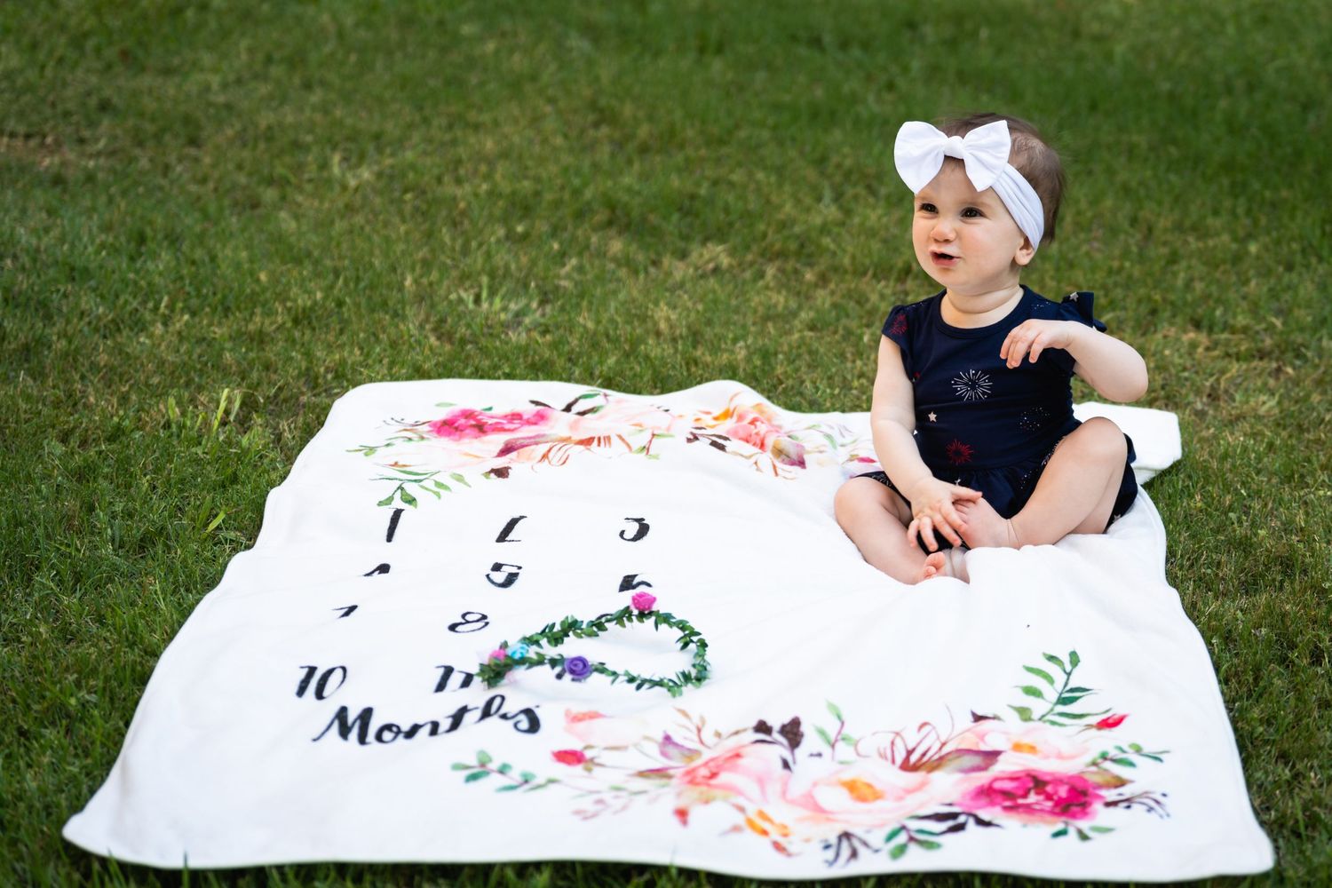 A small child sits on a floral milestone blanket in the grass wearing a bow headband.