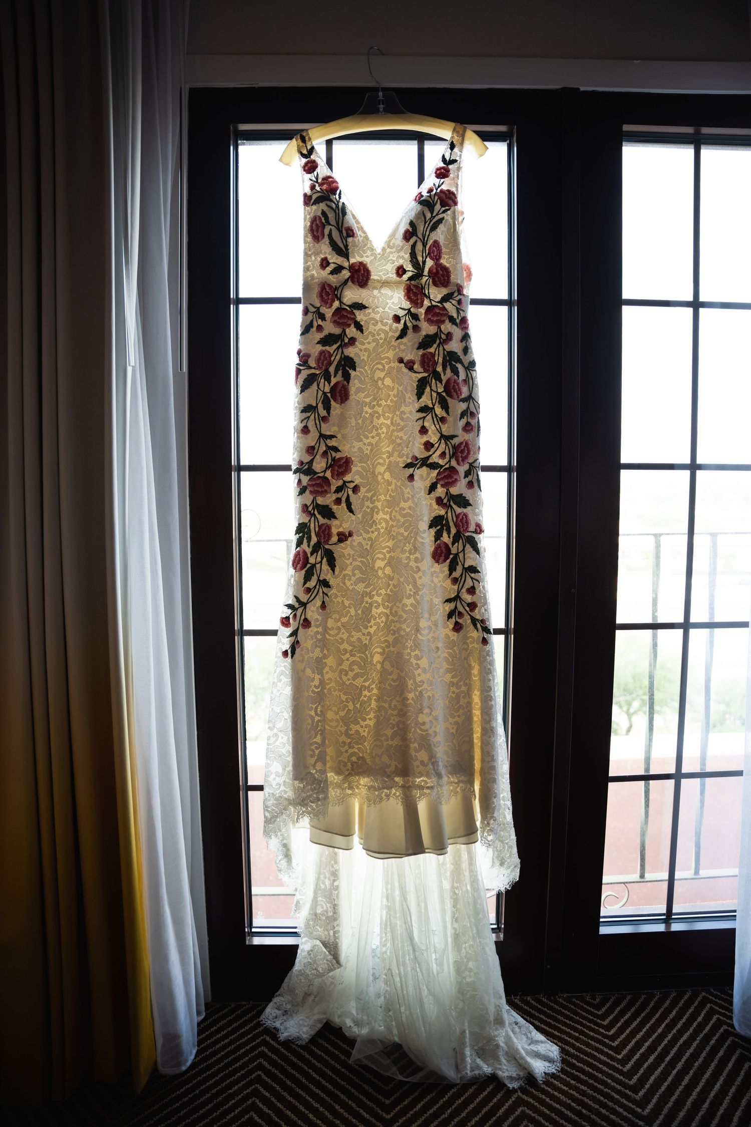 A delicate white wedding dress with floral embellishments hangs elegantly in front of a window.
