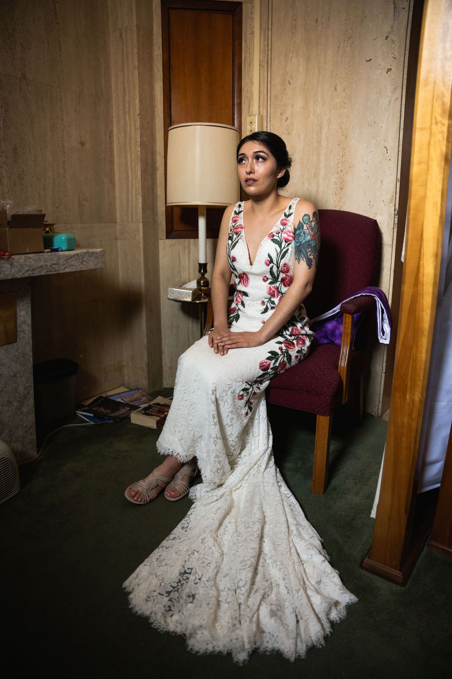 Person seated in vintage chair wearing white lace gown and floral top in dimly lit room.