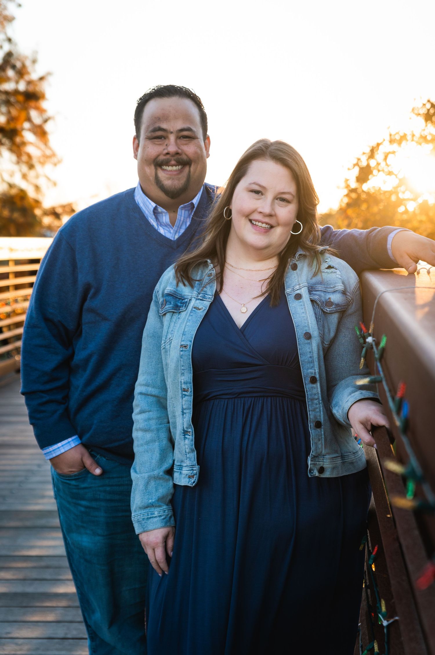 A couple poses on a wooden bridge at sunset, with her wearing a denim jacket and navy dress and him in a blue sweater.