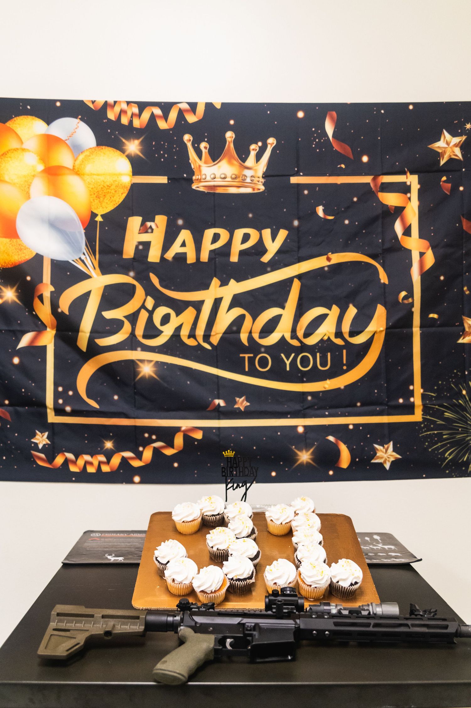 Birthday celebration setup with cupcakes and firearms against a dark backdrop with gold balloons and crown decoration.
