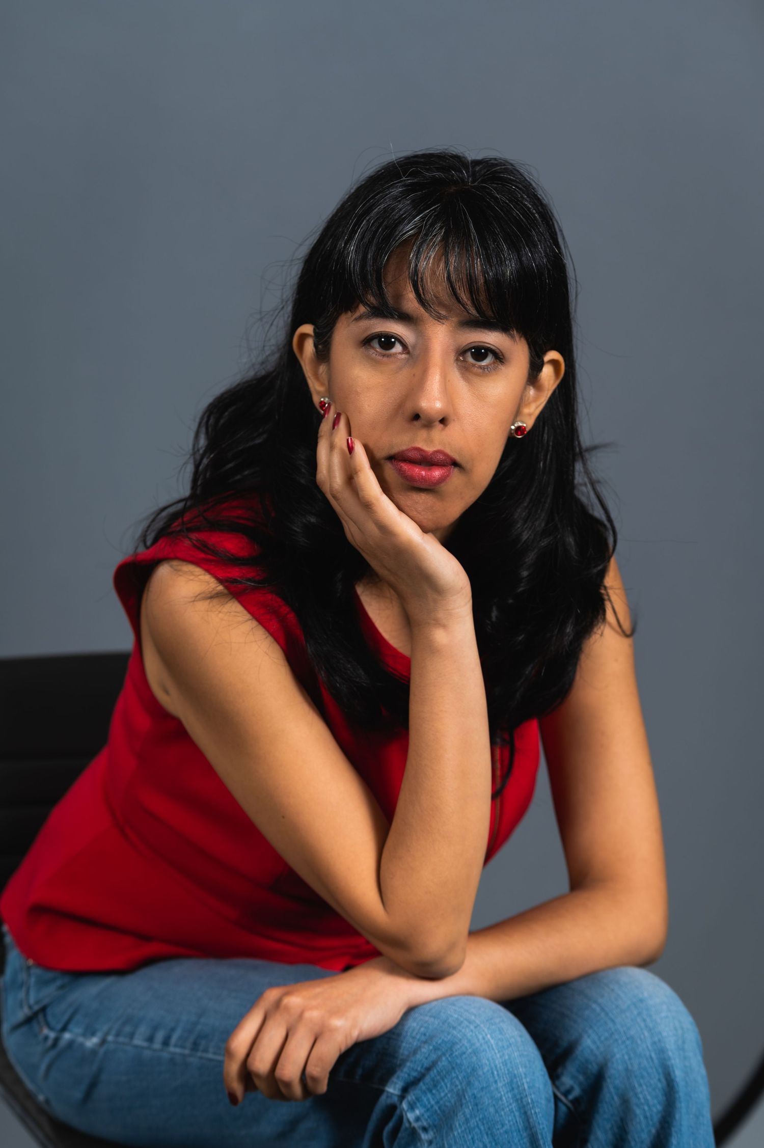 A woman in a red top and jeans poses casually against a grey background.