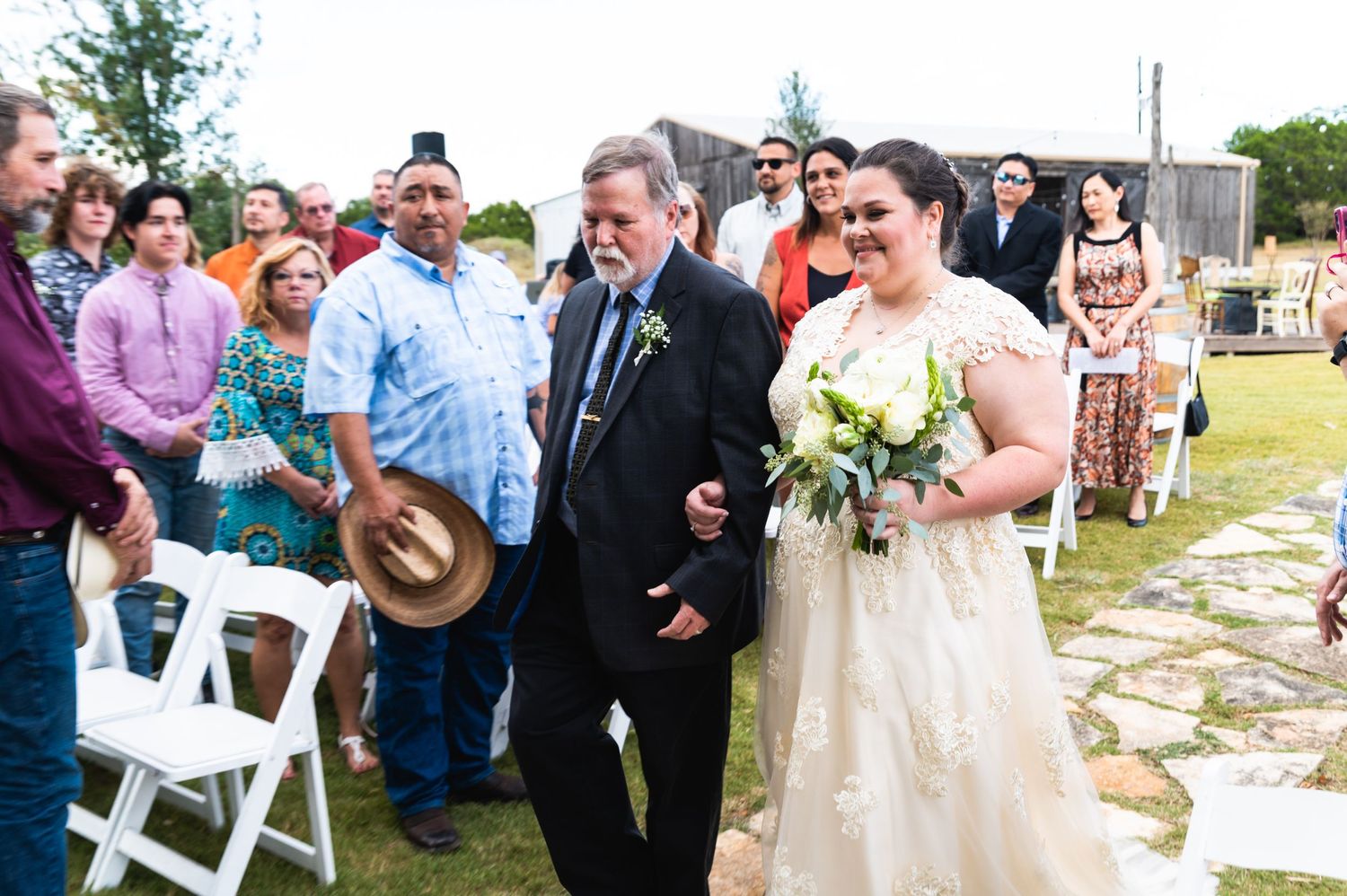 Wedding guests and attendees smile during an outdoor ceremony on a stone pathway.