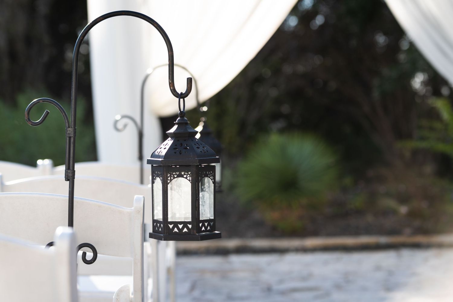 Black decorative lantern hangs from a curved metal hook with white fabric draped in background.