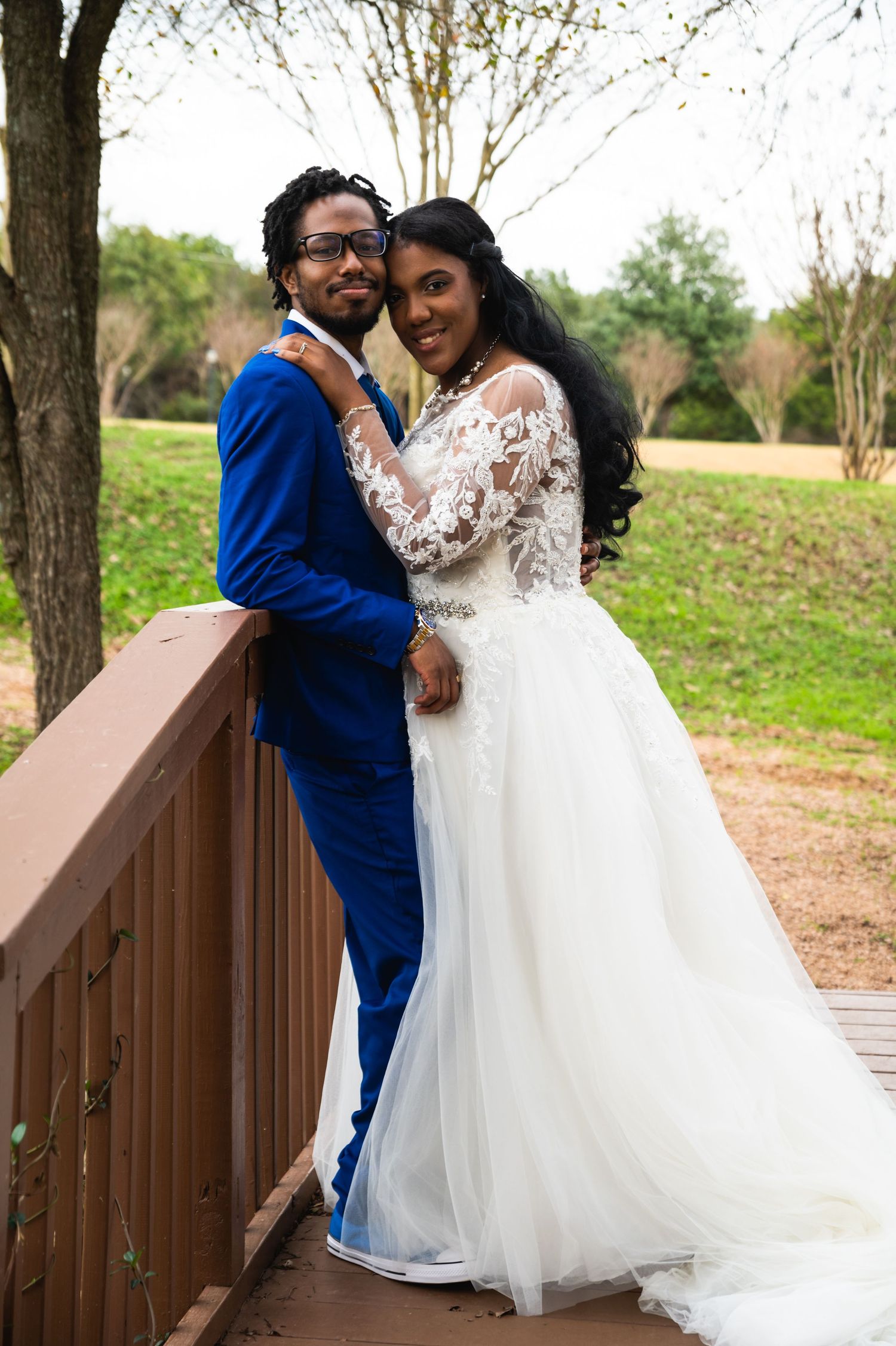 Joyful wedding couple poses on wooden bridge with bride in lace gown and groom in bright blue suit.