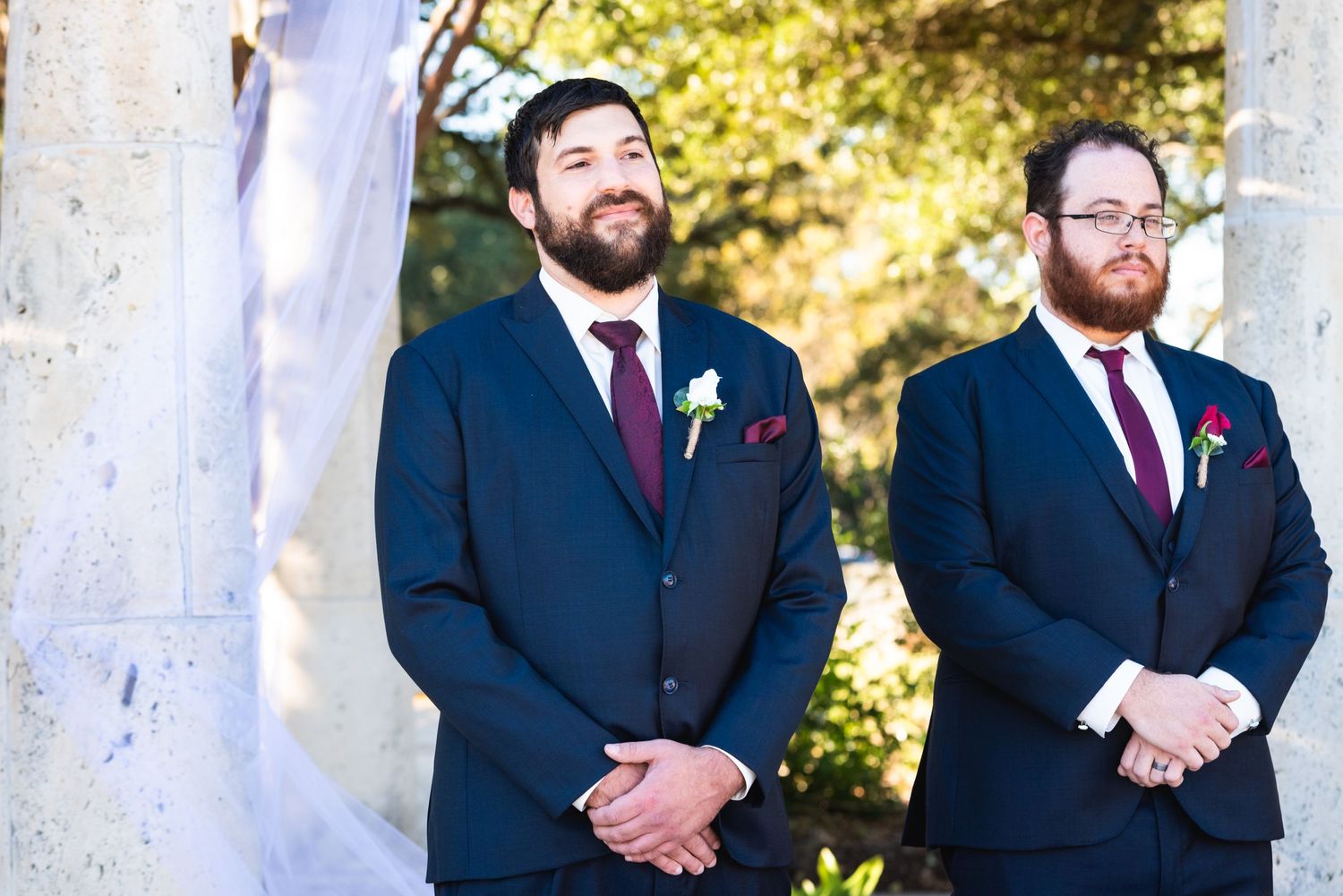 Two men in navy suits with burgundy ties wait at an outdoor wedding altar.