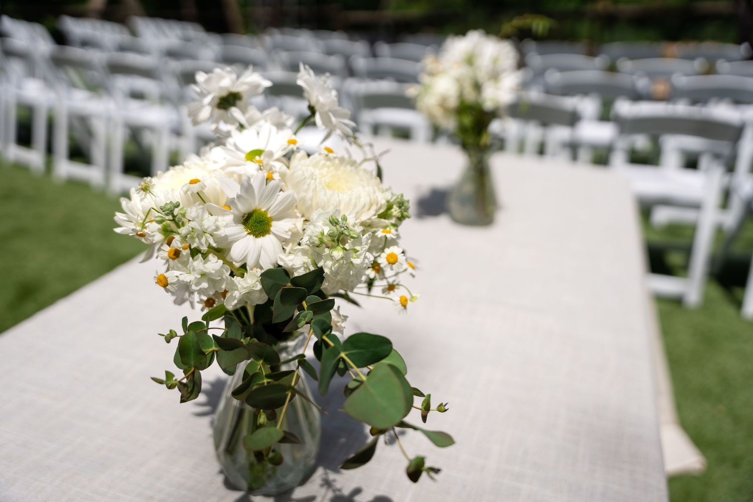 White floral arrangements in mason jars line an outdoor wedding aisle with white chairs.