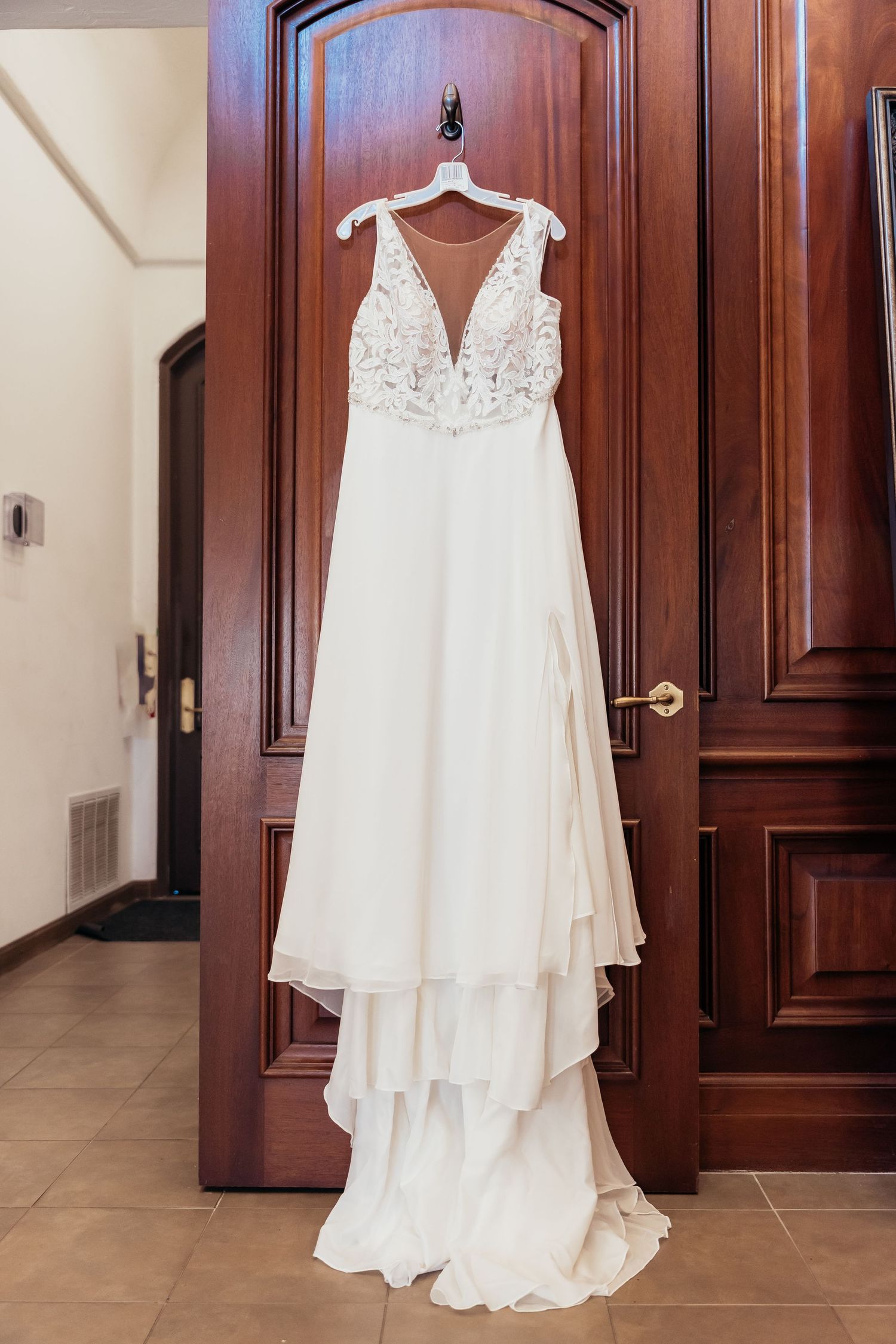 Elegant white wedding dress with lace bodice and flowing chiffon skirt hanging on wooden door.