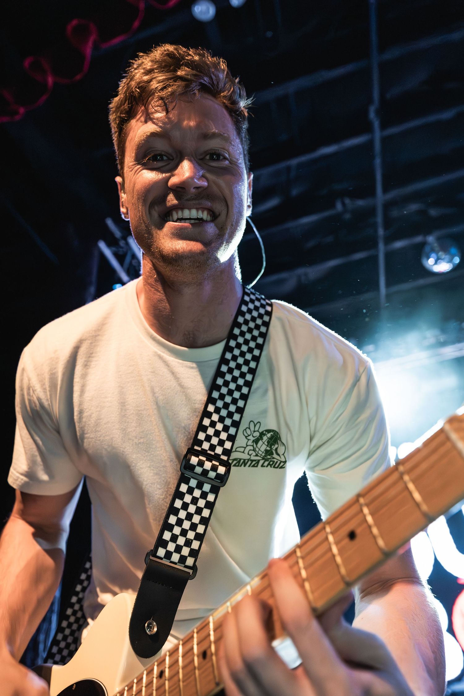 Musician playing electric guitar with a checkered strap performing on stage under bright lights.