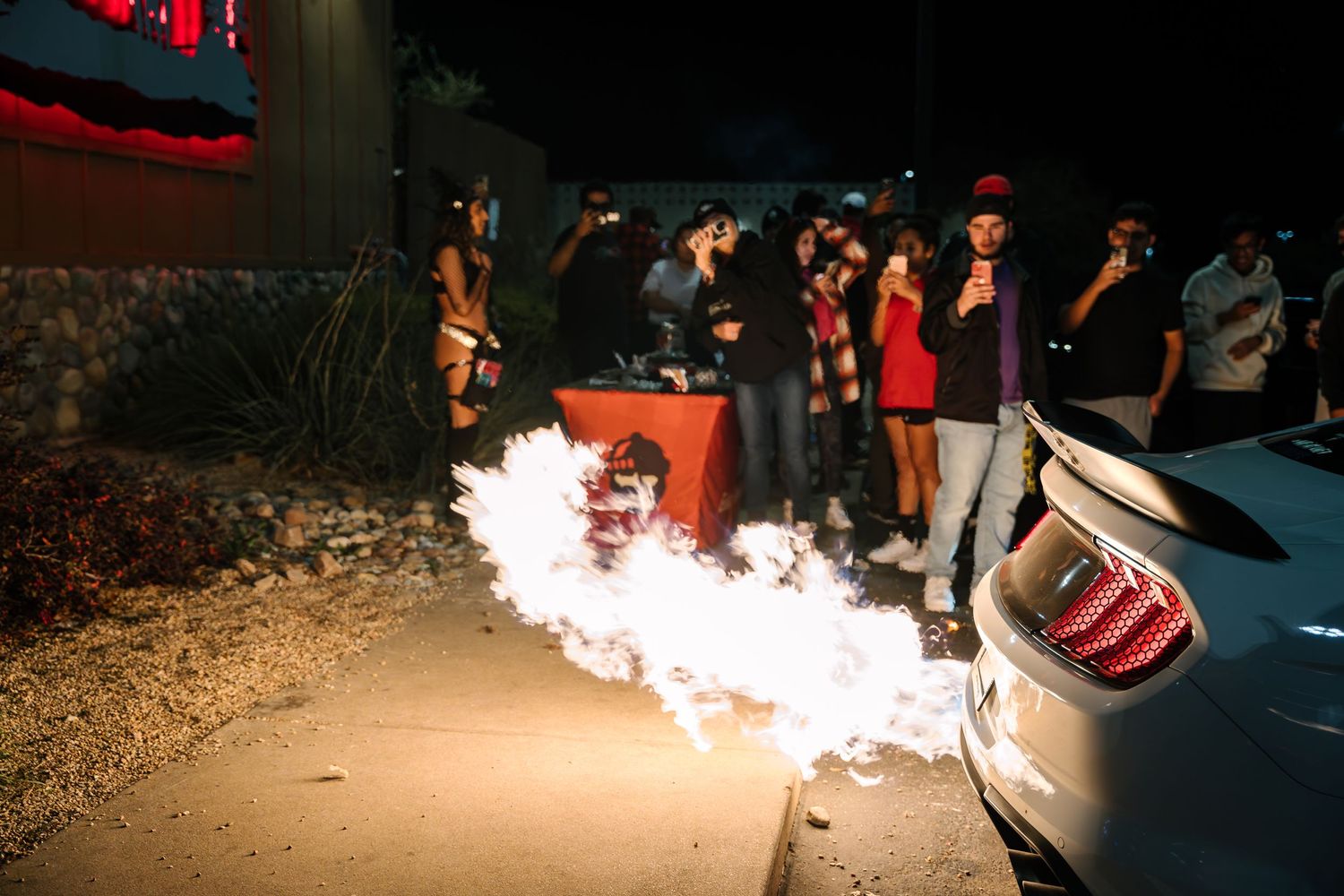 Car enthusiasts gather around vehicle shooting flames from exhaust at night meet.