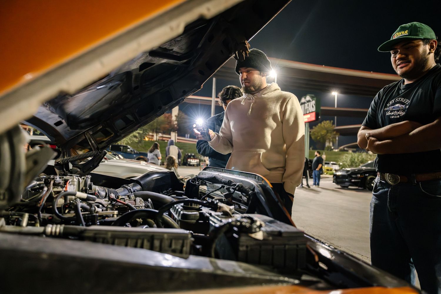 Car enthusiasts examining engine bay of modified vehicle at nighttime car meet.