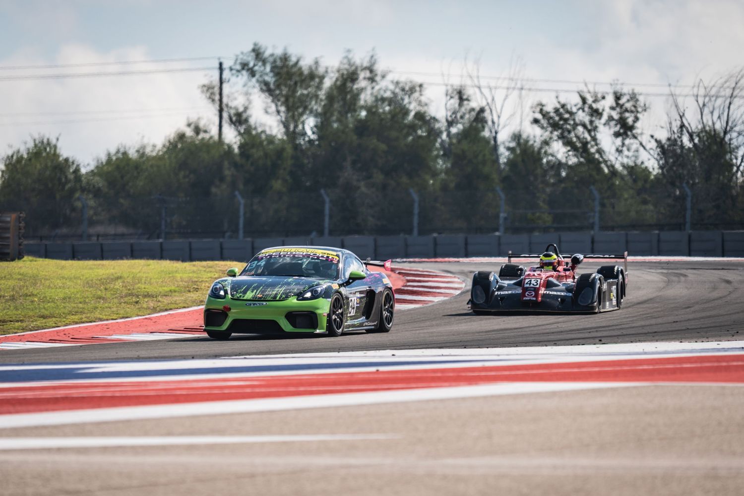 Green sports car leads a race car through a curved section of track with red and white barriers.