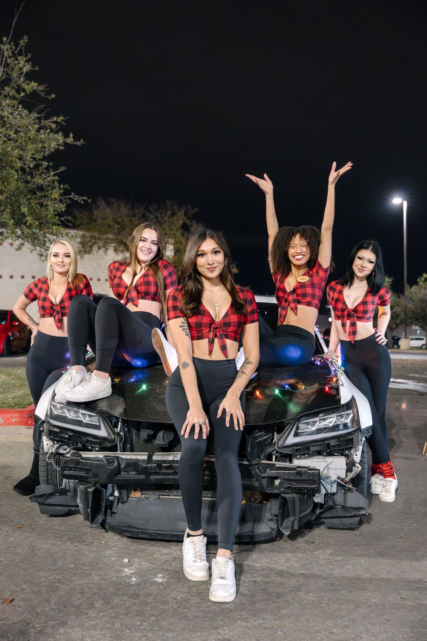 Group in coordinated red plaid tops and dark pants pose on damaged car hood in parking lot.