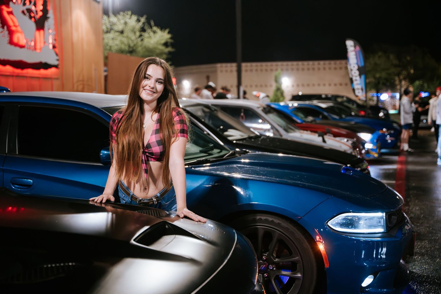 Person in plaid top leaning against blue sports car at nighttime car meetup.