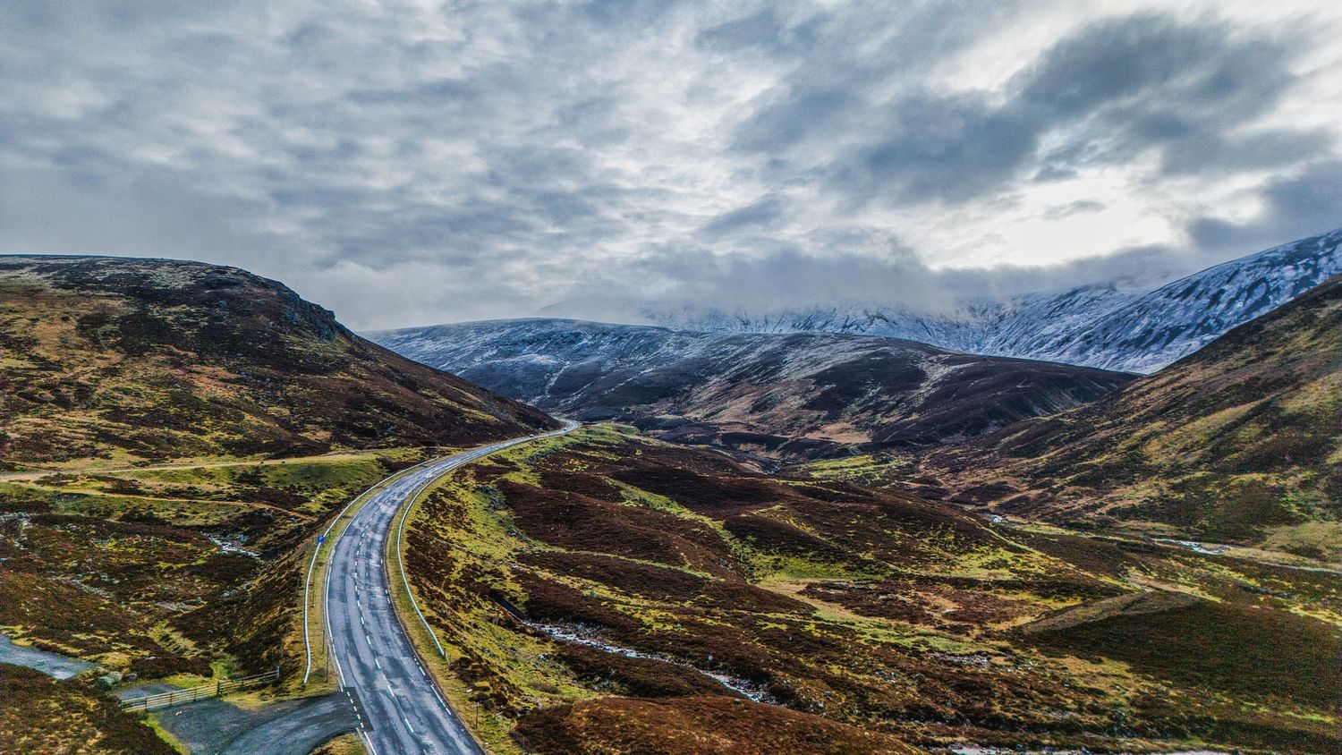 Winding road through Scottish highlands with snow-capped mountains and dramatic cloudy sky.