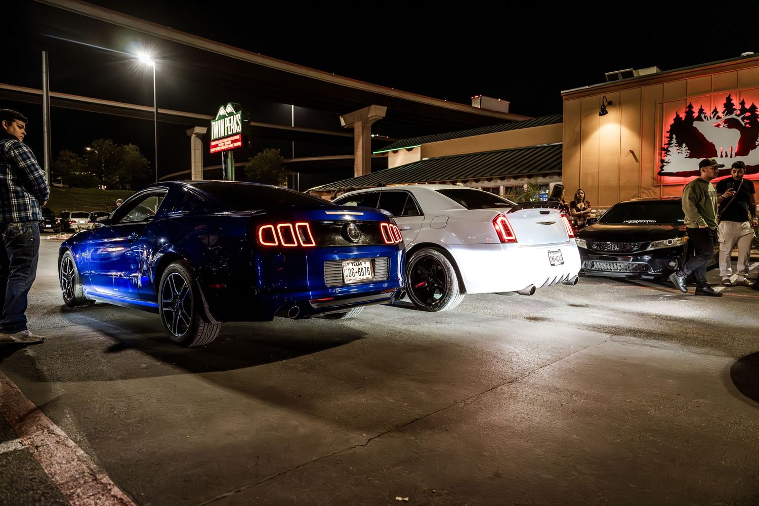 Two sports cars, a blue and white model, parked outside at night under bright lights in a parking lot.
