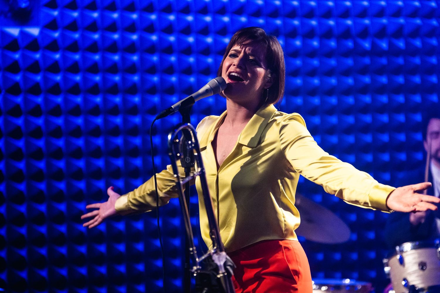 A performer in bright yellow and red sings passionately on stage with a blue textured backdrop.