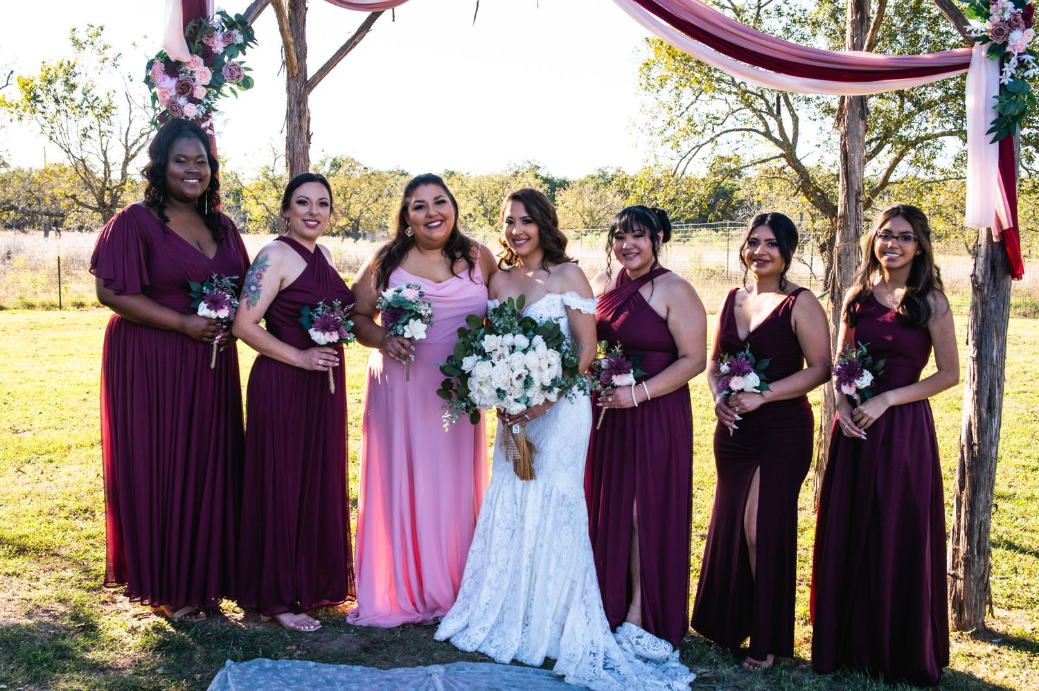 Wedding party poses in deep burgundy and pink dresses under a wooden arch at an outdoor ceremony.