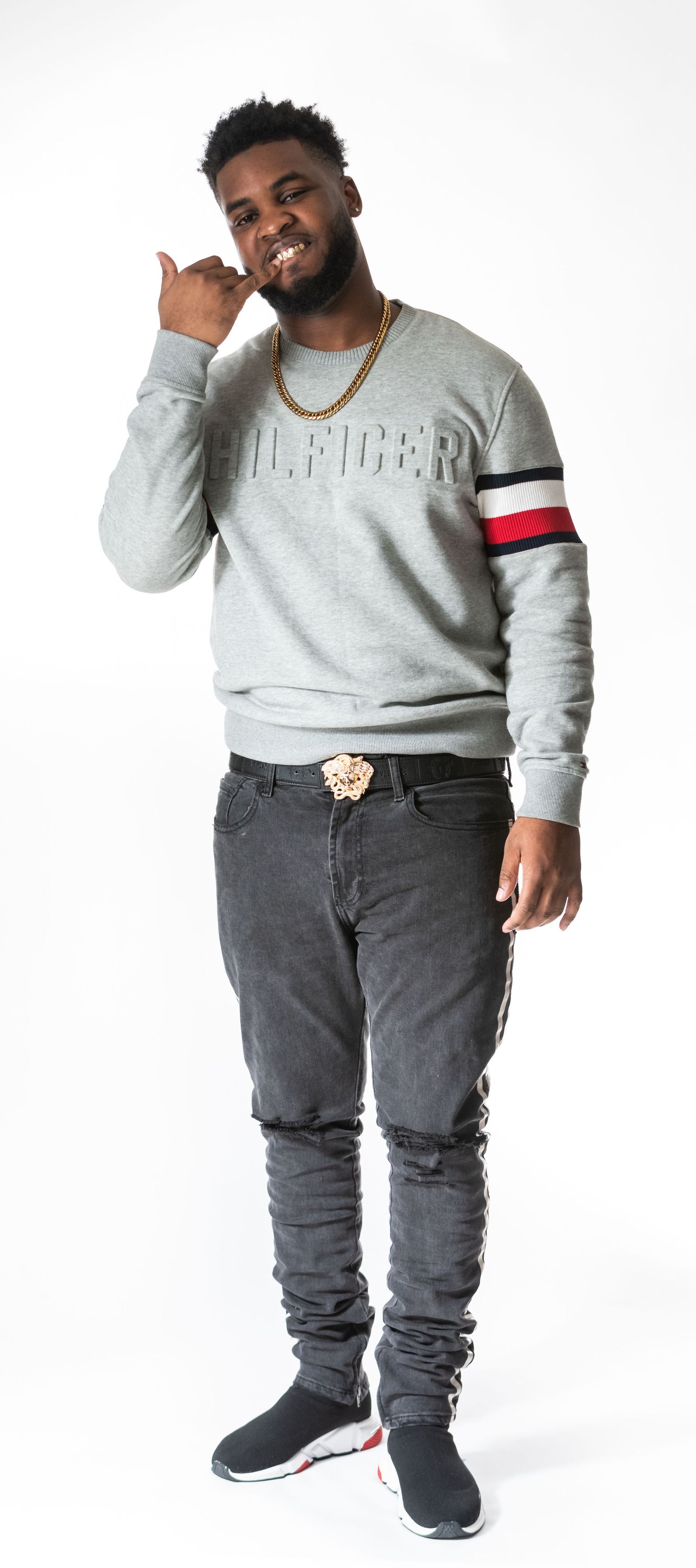 Athletic figure in gray Tommy Hilfiger sweatshirt with striped sleeve detail and distressed jeans poses casually against white backdrop.