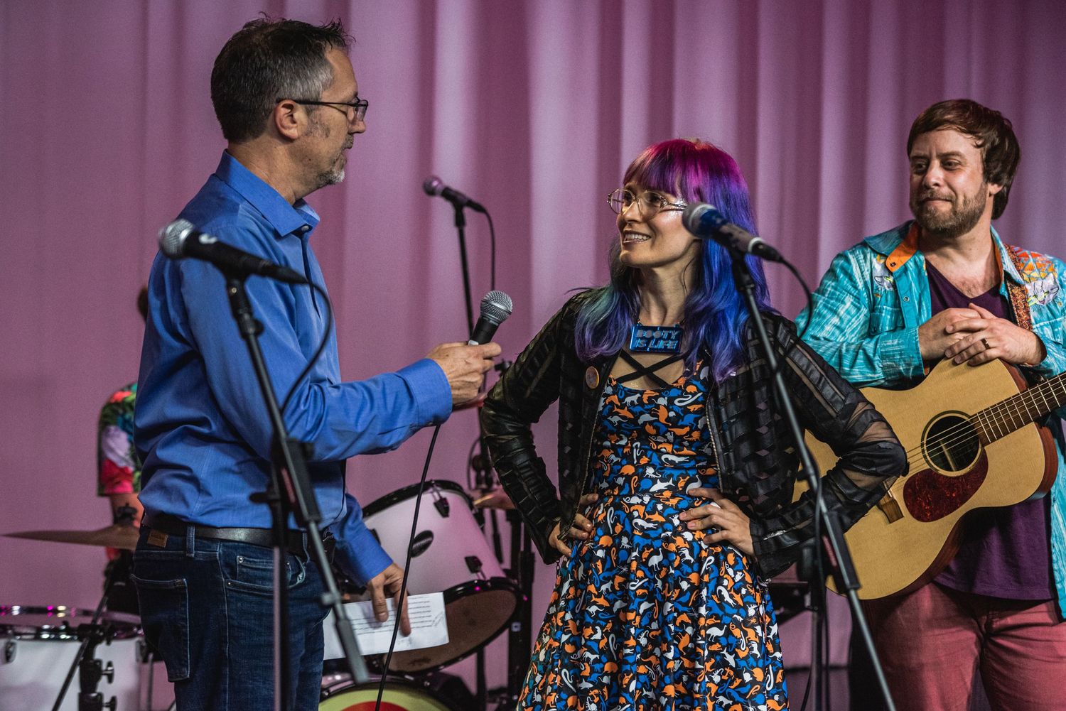 Musicians being interviewed on stage with acoustic guitar and pink curtain backdrop.