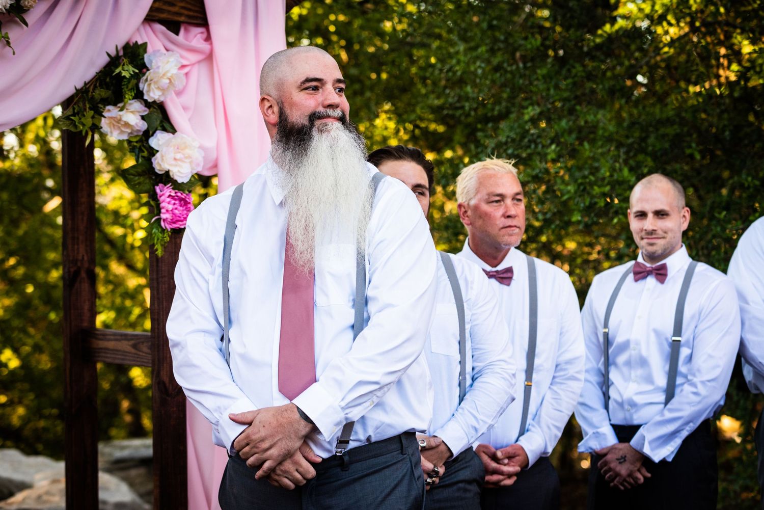 Groomsmen in white shirts and suspenders stand during an outdoor wedding ceremony.